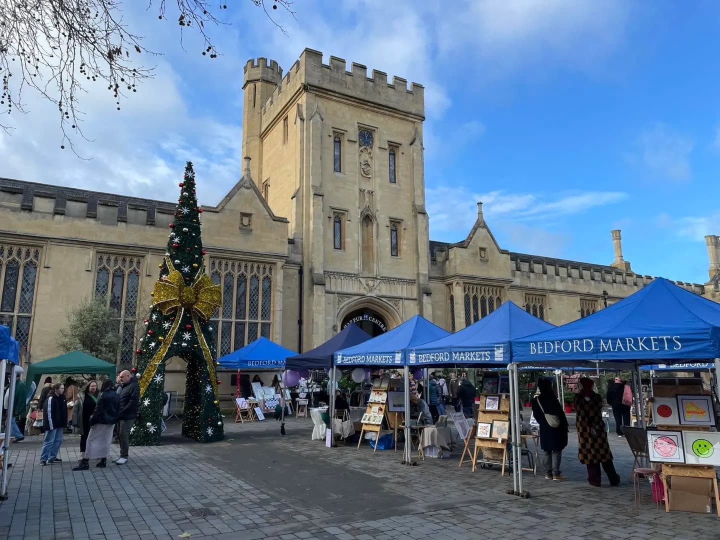 A large decorated Christmas tree with a golden bow stands to the left of several blue vendor tents labeled "Bedford Markets" in front of a grand stone building under a partly cloudy sky.