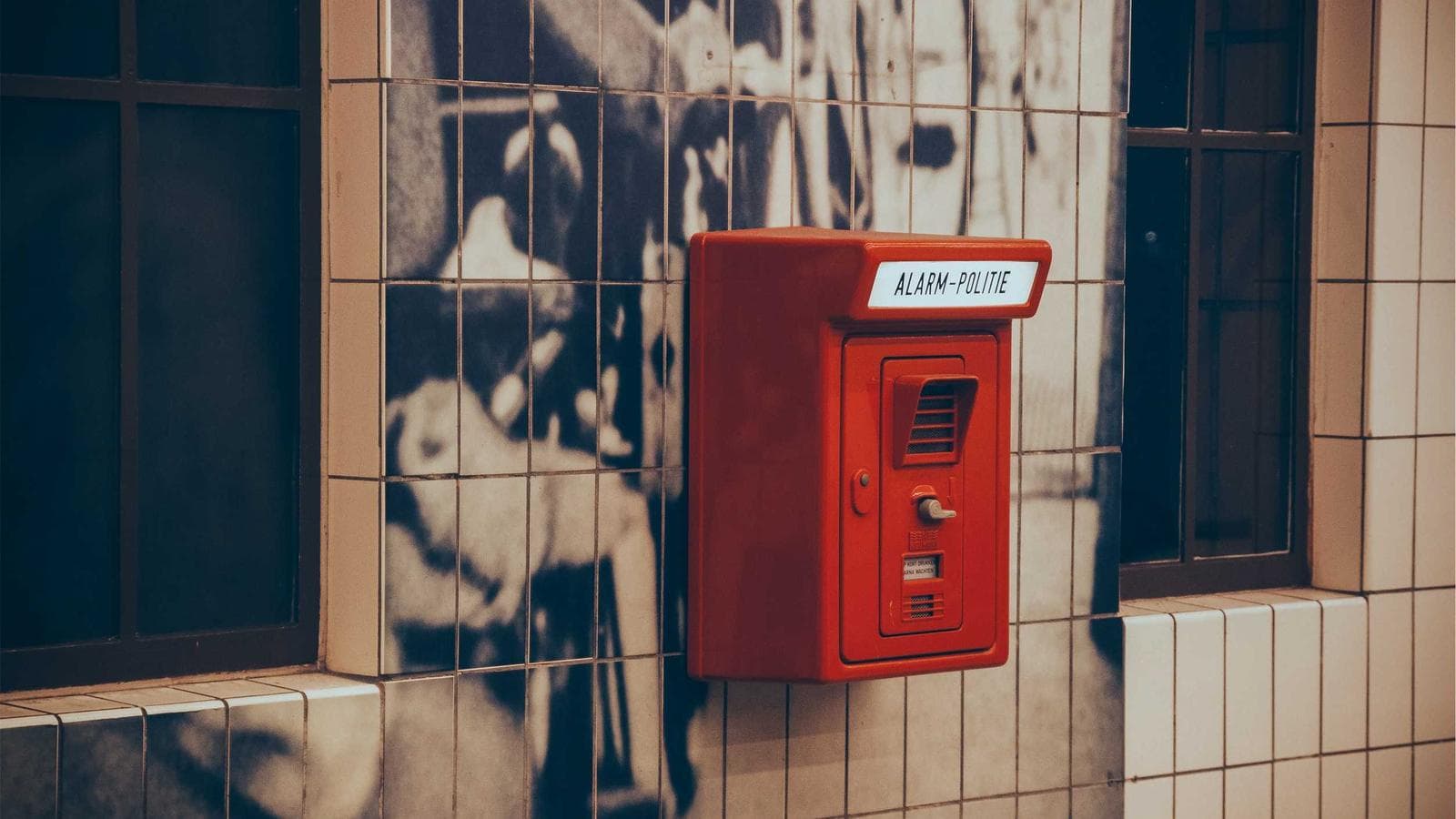 A red alarm box labeled "ALARM-POLITIE" is mounted on a tiled wall with a faint, ghostly image reflecting on some of the tiles.