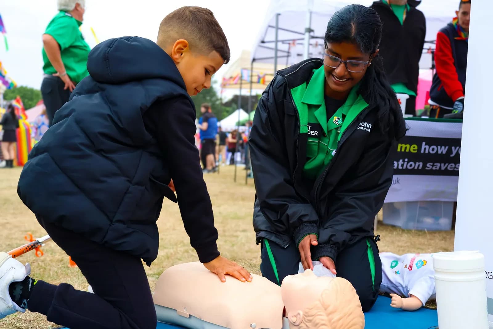 A boy in a black puffer vest kneels next to a smiling instructor in a green and black uniform, both practicing CPR on a mannequin.