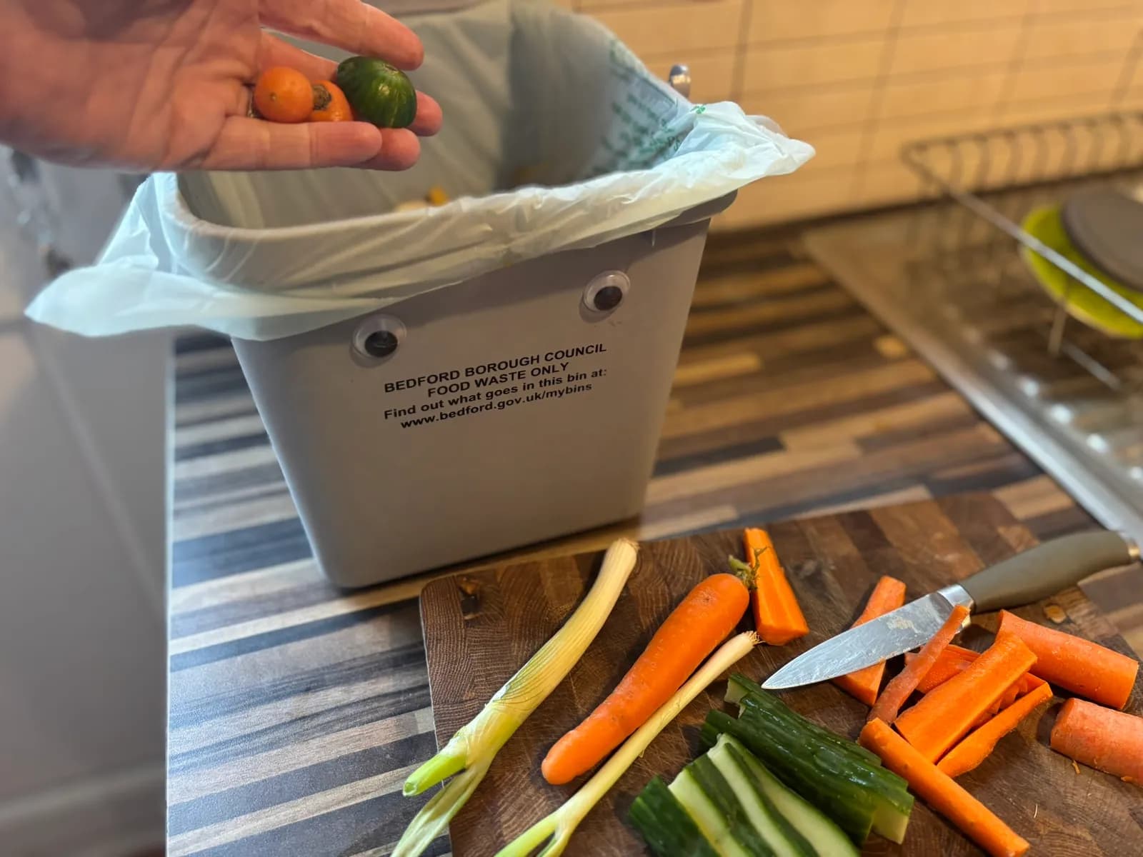 A hand holds small oranges and a green squash over a grey food waste bin, with chopped carrots, leeks, and cucumber on a wooden cutting board in the foreground.